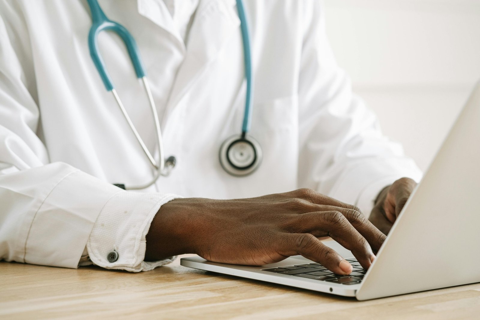 Close-up of a doctor using a laptop, wearing a stethoscope and white coat.